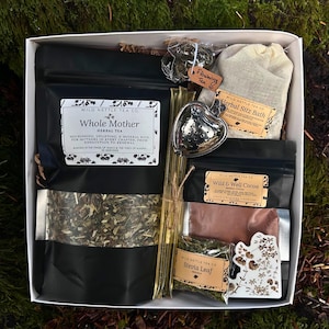 以下が含まれることがあります： お茶とハーブ製品が入った白い箱。箱には「Whole Mother」と「Wild & Well Cocoa」と書かれた黒い袋、ハート型の茶こし、そして「Herbal Sitz Bath」と書かれた小さな袋が入っています。箱は苔むした表面に置かれています。