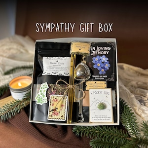 May include: A sympathy gift box with the text "SYMPATHY GIFT BOX". The box contains tea, cocoa, a tea infuser, a candle, and a card that says "A POCKET HUG". The box is decorated with a bird sticker and sprigs of greenery.