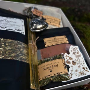 以下が含まれることがあります： 様々なお茶製品が入った白いギフトボックス。ボックスには、黒いお茶の袋、ハート型の金属製のオブジェクト、ハニースティック、花柄の装飾的なセラミック片が含まれています。お茶のパッケージにはテキストが記載されたラベルが付いています。