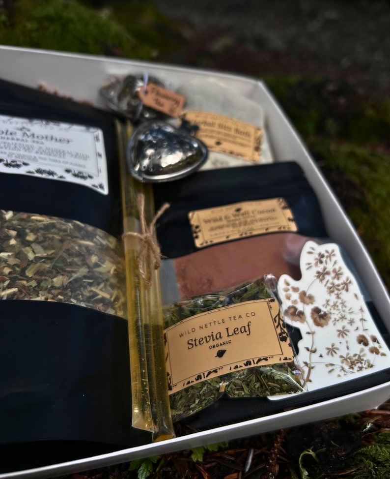 以下が含まれることがあります： 様々なお茶製品が入った白いギフトボックス。ボックスには、黒いお茶の袋、ハート型の金属製のオブジェクト、ハニースティック、花柄の装飾的なセラミック片が含まれています。お茶のパッケージにはテキストが記載されたラベルが付いています。