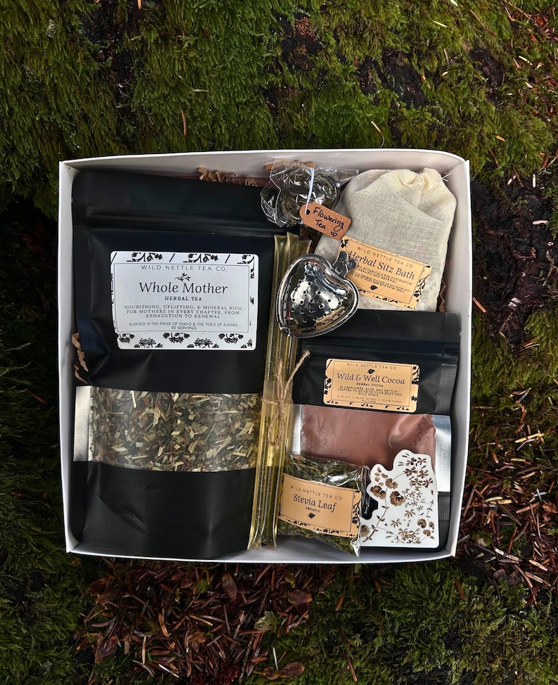 以下が含まれることがあります： お茶とハーブ製品が入った白い箱。箱には「Whole Mother」と「Wild & Well Cocoa」と書かれた黒い袋、ハート型の茶こし、そして「Herbal Sitz Bath」と書かれた小さな袋が入っています。箱は苔むした表面に置かれています。