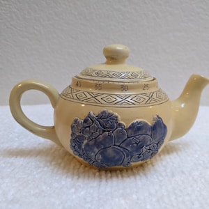 May include: Cream-colored ceramic teapot with a blue fruit design. The lid features a geometric pattern and numbers. The teapot has a curved handle and spout. A decorative kitchenware item.