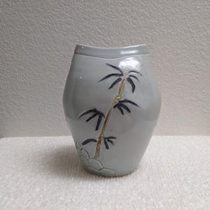 May include: A light gray ceramic vase with a smooth, glossy finish. The front of the vessel features a hand-painted design of a bamboo stalk in shades of brown and black, with a subtle embossed pattern of rounded shapes near the base.