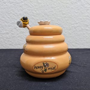 May include: A ceramic honey pot in the shape of a beehive, with a matching lid and a honey dipper topped with a bee. The pot is light orange with the words "honey miel" and a flower design. A charming kitchen accessory.