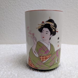 Puede incluir: Taza de cer&aacute;mica blanca con un dise&ntilde;o de geisha. La geisha lleva un kimono verde y sostiene flores rosas. La taza tiene un borde rojo y un dise&ntilde;o decorativo. El pelo de la geisha es negro y est&aacute; adornado con horquillas.