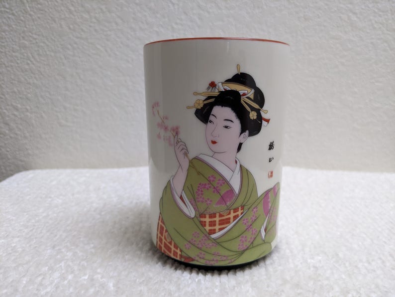Puede incluir: Taza de cer&aacute;mica blanca con un dise&ntilde;o de geisha. La geisha lleva un kimono verde y sostiene flores rosas. La taza tiene un borde rojo y un dise&ntilde;o decorativo. El pelo de la geisha es negro y est&aacute; adornado con horquillas.