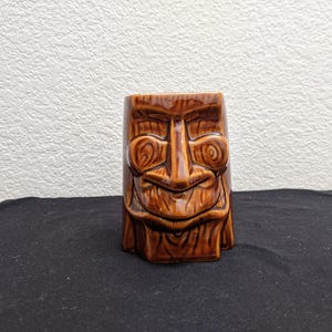 May include: A brown ceramic tiki mug with a carved face design. The mug features a detailed depiction of a tiki god with a wide smile, large eyes, and textured wood-like details. The mug is a square shape.