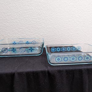 May include: Two rectangular glass baking dishes with a light blue trim and decorative floral patterns in blue and green. The dishes are sitting on a black surface.