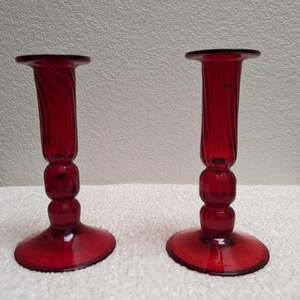 May include: Two red glass candle holders. Each has a round base, a series of rounded sections, and a tall, fluted top. The glass has a glossy finish. The candle holders are displayed on a white surface.