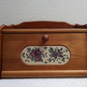 May include: A wooden bread box with a medium brown finish, featuring a curved top edge and a central oval ceramic inlay. The inlay displays a colorful illustration of red apples and purple grapes with green leaves. A small round wooden knob is centered.