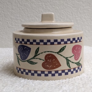 May include: A cream-colored ceramic sugar bowl with a lid, featuring a horizontal vine pattern with purple, brown, and pink heart-shaped leaves. The top and bottom edges are decorated with a blue and white checkered border pattern.