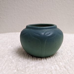 May include: A small, round, matte teal ceramic bowl featuring a subtle, embossed leaf pattern around the upper rim. The vessel has a smooth, curved silhouette with a wide opening and a flat base, showcasing a consistent, muted blue-green color.