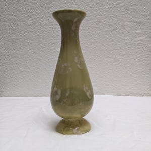 May include: A green ceramic vase with a flared top and a rounded body. The vase has a pedestal base and is decorated with a subtle floral pattern. The vase is approximately 8 inches tall.