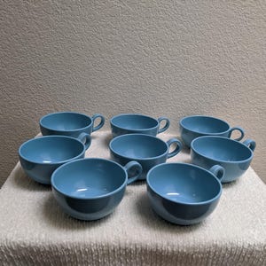May include: A set of eight light blue ceramic soup cups with handles. The cups are arranged on a white textured surface, showcasing their rounded shape and simple design. The cups are a solid, matte color.