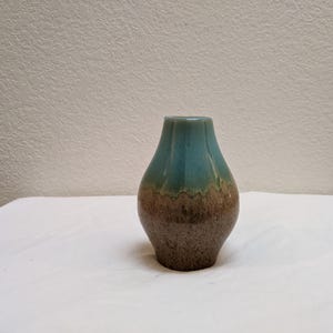 May include: A small, handcrafted ceramic vase with a unique color gradient. The top of the vase is a vibrant teal, transitioning to a brown and tan base. The vase has a textured surface and a fluted rim, adding to its artisanal appeal.