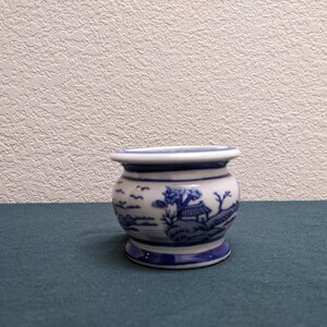May include: A small, round, ceramic pot with a blue and white design. The pot features a landscape scene with a house, trees, and birds. The rim and base are blue, and the pot sits on a dark teal surface.