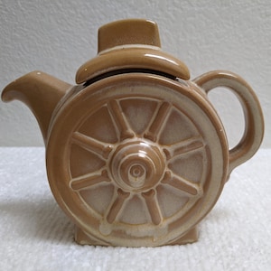 May include: A tan ceramic teapot with a wagon wheel design on the side. The teapot has a handle, a spout, and a lid. The wheel design is detailed with spokes and a central hub. The teapot is a light brown color.