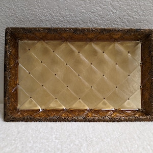 May include: A rectangular glass tray featuring a translucent amber center with a diamond-quilted pattern and small dark dots at each intersection. The tray is framed by a wide, textured border in a deep bronze tone with a repeating shell motif.