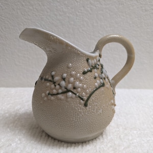 May include: A beige ceramic creamer jug with a textured, pebbled surface. The jug features a raised, hand-painted design of dark green branches with small, white, rounded floral buds. It has a curved handle and a wide pouring spout.