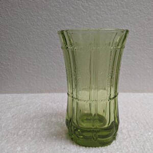 May include: A green glass vase with a textured design. The vase has a flared top and a detailed pattern of vertical lines and rope-like accents. The vase is empty and sits on a white surface.