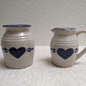May include: A pair of speckled off-white ceramic vessels, including a small pitcher with a handle and a matching open-top jar. Both pieces feature a hand-painted dark blue heart motif on the front, accented by small blue leaf-like shapes.