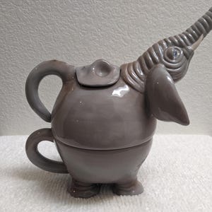 May include: A glossy, light gray ceramic teapot shaped like an elephant. The elephant's trunk forms the spout, while its ear and head create the lid. A curved handle extends from the back of the round body, which sits on four short, sturdy legs.
