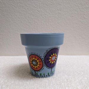 May include: A small, light blue painted flower pot with a decorative floral design. The pot features two large, stylized flower designs in orange, purple, and yellow. The rim of the pot is a darker shade of blue.