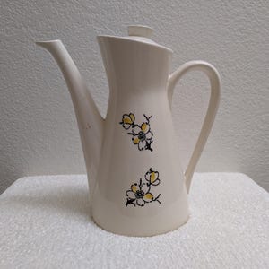 May include: A cream-colored ceramic coffee pot featuring a tapered body, a curved handle, and a matching lid. The exterior is decorated with two floral motifs consisting of black outlines and yellow centers on the front of the vessel.