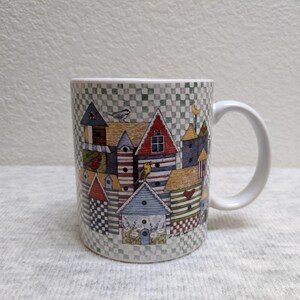 May include: A ceramic coffee mug featuring a whimsical village scene with colorful houses, birdhouses, and birds. The mug has a white handle and a checkered pattern at the top and bottom. The design includes red, blue, and yellow accents.