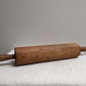 May include: A cylindrical wooden rolling pin with a smooth, medium-brown finish. The tool features a central barrel section for flattening dough and two rounded, tapered handles on each end, showing natural wood grain and minor surface wear.