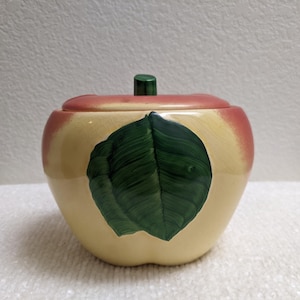 May include: A ceramic apple-shaped jar with a removable lid. The jar features a cream-colored base with a gradient of reddish-pink around the top rim. A large, dark green painted leaf is centered on the front, and a small green stem sits on the lid.