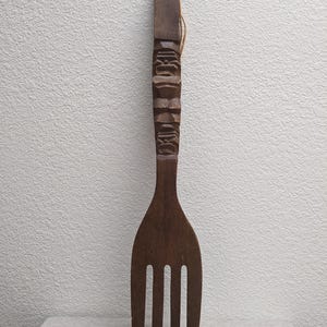 May include: A large, dark brown wooden fork with a carved handle. The handle features a series of carved faces and geometric shapes. A natural fiber cord is attached at the top. The fork has four tines.
