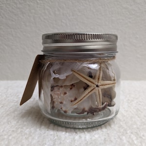 May include: A clear glass mason jar with a silver metal lid filled with various seashells and a light tan starfish. A piece of brown twine is wrapped around the neck of the jar, securing a blank brown rectangular paper tag to the side.