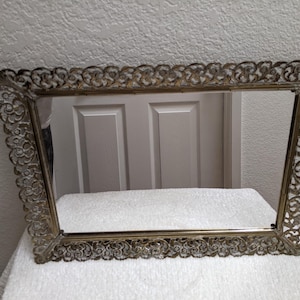 May include: A rectangular mirror featuring an ornate, metallic gold-toned filigree frame with intricate scrollwork and floral-inspired patterns along all four edges. The reflective glass surface is clear and set within the decorative metal border.