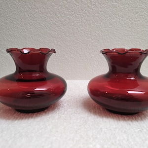 May include: Two matching deep red glass vases with a wide, rounded base and a narrow neck. Each vase features a distinct ruffled, scalloped rim at the top opening. The glass is translucent, allowing light to pass through the rich, dark crimson color.
