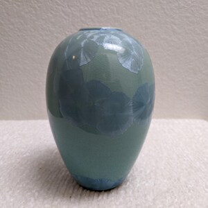 May include: A teal-colored ceramic vase with a rounded shape. The vase features a glossy finish and a pattern of light blue, flower-like designs. The vase is a decorative item.