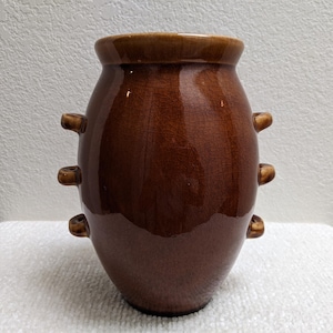 May include: A brown ceramic vase with a glossy finish. The vase has a rounded body with a slightly flared rim and three pairs of small, rounded handles. The color is a rich, warm brown with darker accents.