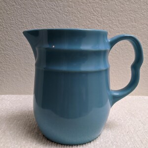May include: A light blue ceramic pitcher with a smooth, glossy finish. The vessel features a rounded body, a wide pouring spout, and a curved handle. The exterior surface is marked by a subtle, horizontal indented band encircling the upper section.
