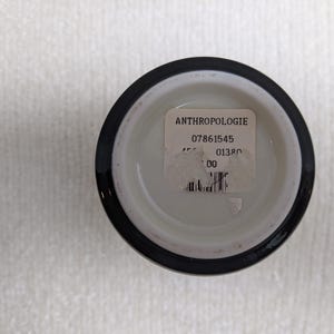 Puede incluir: Un peque&ntilde;o recipiente redondo de cer&aacute;mica blanca con un borde negro. Una etiqueta blanca con la palabra "ANTHROPOLOGIE" y otro texto est&aacute; adherida al fondo interior del recipiente. El recipiente se encuentra sobre una superficie blanca texturizada.