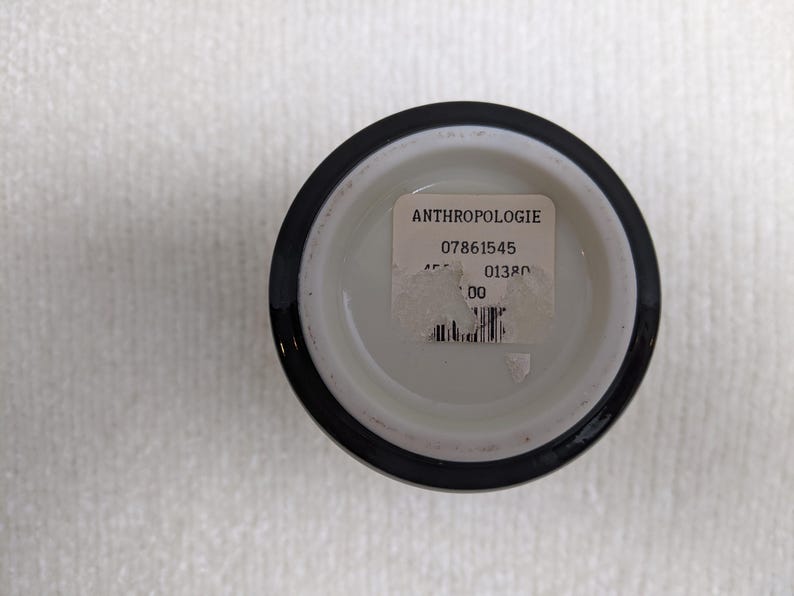 Puede incluir: Un peque&ntilde;o recipiente redondo de cer&aacute;mica blanca con un borde negro. Una etiqueta blanca con la palabra "ANTHROPOLOGIE" y otro texto est&aacute; adherida al fondo interior del recipiente. El recipiente se encuentra sobre una superficie blanca texturizada.