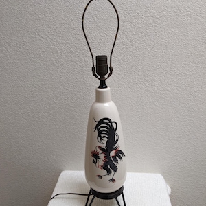 May include: Cream-colored ceramic table lamp with a black and red rooster design. The lamp has a black metal base and a dark brown metal lamp frame. The lamp is on a white surface.