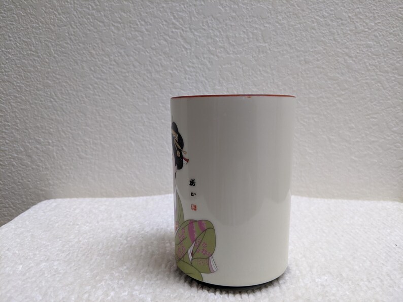 Puede incluir: Taza de cer&aacute;mica blanca con borde rojo, con un dise&ntilde;o de geisha japonesa. La geisha est&aacute; representada con vestimenta tradicional, peinado detallado y kimono colorido. La taza es cil&iacute;ndrica.