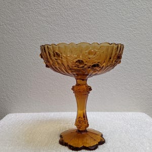 May include: Amber-colored, vintage glass pedestal bowl. The bowl has a scalloped rim and is adorned with a floral pattern. The stem and base are also detailed with decorative elements. The bowl is likely used for serving desserts or displaying decorative items.
