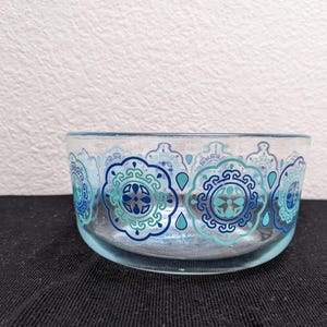 May include: A clear glass bowl with a light blue tint. The bowl is decorated with a repeating pattern of blue and teal floral designs. The bowl sits on a black surface against a white background.