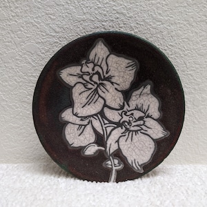 May include: A round, ceramic coaster with a dark brown background and a white orchid flower design. The orchid has detailed petals and a stem. The coaster is likely used for placing drinks on a table to protect the surface.