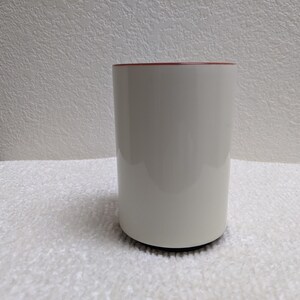 Puede incluir: Una taza de cer&aacute;mica blanca con interior rojo. La taza cil&iacute;ndrica est&aacute; sobre una superficie blanca y tiene una base negra. La taza est&aacute; vac&iacute;a y parece limpia.
