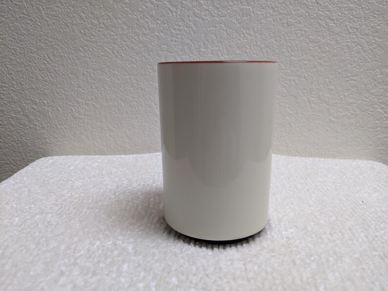 Puede incluir: Una taza de cer&aacute;mica blanca con interior rojo. La taza cil&iacute;ndrica est&aacute; sobre una superficie blanca y tiene una base negra. La taza est&aacute; vac&iacute;a y parece limpia.