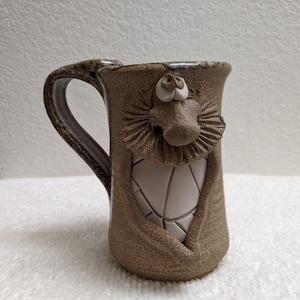 May include: A handmade ceramic mug with a speckled brown finish and a large curved handle. The front features a sculpted, light-colored face with a ruffled collar and a textured, geometric pattern on the lower body, creating a unique character design.