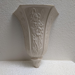 May include: Cream-colored wall vase with a floral iris design. The vase has a flared top and a gold trim. The iris design is raised and detailed. The vase is mounted on a wall.