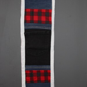 May include: A rectangular wall hanging made from repurposed denim and plaid fabric. The design features sections of blue denim, red and black plaid, and a black denim pocket, all framed with a white border. The piece is designed to be hung on a wall.